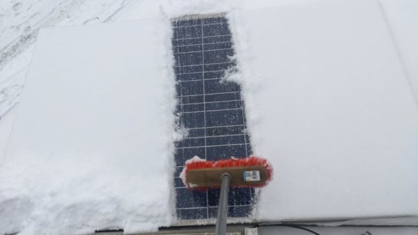 Ein polykristallines Solarmodul ist fast unter Schnee begraben. Ein Besen kehrt es gerade frei.